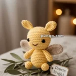 A cute yellow crocheted firefly toy with black eyes and brown wings, placed on a surface decorated with green leaves. A tag next to it reads "Dream bright." Soft, warm lighting and a blurred background create a cozy atmosphere.