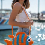 A woman wearing a white tank top and denim shorts smiles while carrying a colorful fish-shaped crocheted bag with orange and blue stripes, standing near a waterfront.