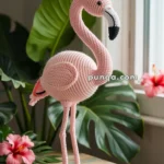 A crocheted pink flamingo standing on a wooden table, surrounded by green leaves and red hibiscus flowers near a window.