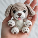 A small crocheted dog toy with brown and white details and a pink tongue, held in a person's hand against a white background.