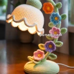 A crochet lamp with a scalloped shade and base, featuring a design of intertwined stems adorned with colorful crochet flowers in orange, blue, pink, and purple. The lamp is lit, highlighting the handmade detail, and sits on a wooden table near a curtain in the background.