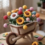 A crochet arrangement of colorful flowers, including sunflowers and round blooms, placed in a handmade crochet wheelbarrow on a wooden table with scattered leaves and flowers.