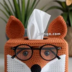 A crocheted tissue box cover shaped like a fox wearing glasses, with tissues coming out from the top, set in a background with a green plant and a sunflower decoration.
