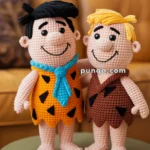 Two crocheted dolls resembling cartoon characters, one with black hair and an orange outfit with a blue tie, and the other with blond hair and a brown outfit, standing on a small round table in a room with a brown couch in the background.