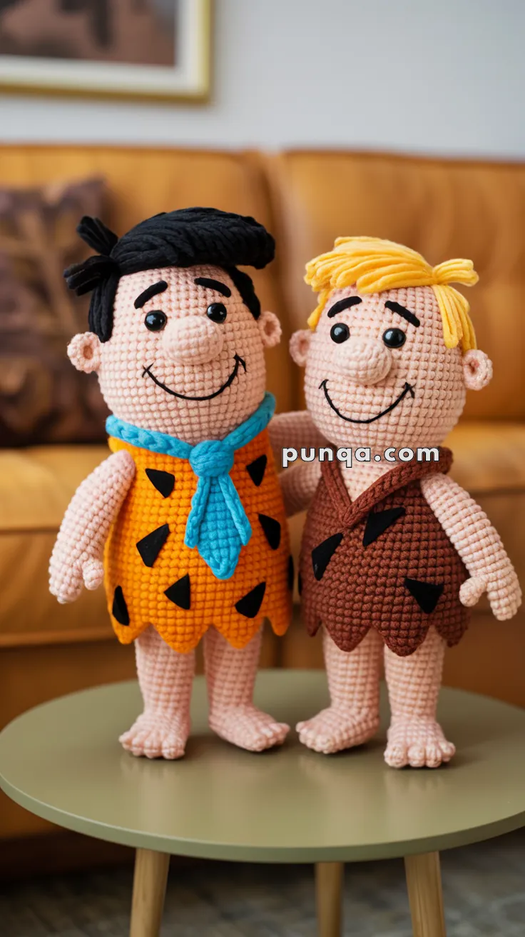 Two crocheted dolls resembling cartoon characters, one with black hair and an orange outfit with a blue tie, and the other with blond hair and a brown outfit, standing on a small round table in a room with a brown couch in the background.