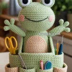 A crochet frog-shaped organizer with multiple pockets holding scissors, pens, and other stationery items, placed on a table in a cozy room setting.
