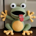 A crocheted frog toy with large, googly eyes, a wide open mouth, a red tongue sticking out, and yellow hands and feet with four toes. The frog's body is green with a yellow belly. It is sitting on a wooden floor against a beige door.