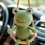 A crocheted green frog hanging from a string inside a car, with the dashboard visible in the background.