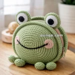 A round, crocheted bag shaped like a cute green frog with big eyes, sitting on a wooden table next to a potted plant.