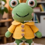 A crocheted frog toy with large eyes and a smile, wearing a yellow jacket with buttons and matching yellow shoes, stands on a plush surface with a cozy room setting in the background.