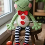 A crocheted green frog with large eyes, a red bow tie, and hearts on its belly, wearing black and white striped legs and red shoes, sitting on a wooden stool next to small knitted mushrooms and leaves.