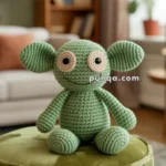 A green crocheted stuffed froglet toy with large eyes and round ears sitting on a green plush ottoman in a cozy living room setting.