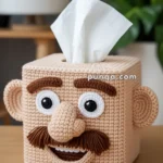 A crochet tissue box cover shaped like a human face with prominent features including large white eyes, a thick mustache, and a large nose, situated on a wooden surface with a white tissue protruding from the top.