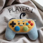 A crocheted video game controller with a smiling face design, featuring colorful buttons, resting on a soft fabric with 'PLAYER 1' embroidered on it.