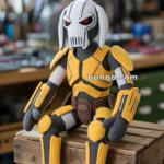 A crocheted figure resembling a robot or armored character sitting on a wooden crate, featuring a gray, yellow, and brown color scheme with a detailed helmet and intricate armor design.