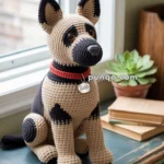 A crocheted dog resembling a German Shepherd is sitting on a wooden table by a window, wearing a red collar with a tag labeled 'Buddy.'
