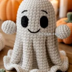 A crocheted ghost with a smiling face sits on a wooden table surrounded by crocheted pumpkins and lit candles in the background.