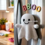 A crocheted white ghost decoration with a smiling face is attached to a refrigerator, beneath magnetic letters spelling 'BOO' in colorful plastic. The kitchen background includes a wooden countertop with plants and kitchen items visible.