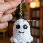 A hand holding a crocheted ghost keychain with large black eyes and a smiling face in a cozy room setting.