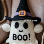 A crochet ghost pillow with a black witch hat featuring a purple band and orange buckle, embroidered with "BOO!" on its body, seated on a vintage armchair.