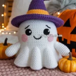 A crocheted ghost toy wearing a purple witch hat with an orange band, placed on a pink textured mat between two small pumpkins, with fairy lights in the background and a carved pumpkin nearby.