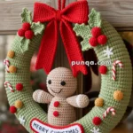 A crocheted Christmas wreath decorated with holly leaves, berries, candy canes, and small baubles, featuring a gingerbread man in the center and a red bow at the top. The wreath has a 'Merry Christmas' sign at the bottom.