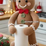 A crocheted gingerbread man holding a roll of paper towels in a cozy kitchen setting, decorated with festive lights and holiday-themed items like gingerbread cookies and candy canes.