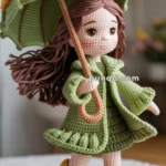 A crocheted doll with brown hair wearing a green coat and holding a green umbrella decorated with flowers, standing on a green base. The doll is also wearing pink boots with yellow accents.