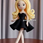 A crocheted doll wearing a black dress and black heels, with long blonde hair and a black bow, standing on a wooden surface with a soft background curtain and light overhead.