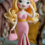 A crocheted doll with long blonde hair, wearing a pink dress with a slit, a large pink bow on its head, and holding a small pink purse. The doll is standing on a wooden surface.