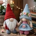 Two crochet gnomes, one with a red hat and white beard, and the other with a blue dress and hat adorned with pink flowers, stand holding hands. A wooden sign reading 'Always & Forever' is placed in front of them, and fairy lights and decorative hearts are in the background.