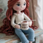 A crocheted doll with long, red hair, wearing a white top and blue pants, sitting on a wooden surface while holding a mug labeled 'Good Vibes.'