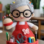 A crocheted doll resembling an elderly woman, with grey hair tied in a bun, large black glasses, and a red apron holding crafting supplies, displayed on a table.