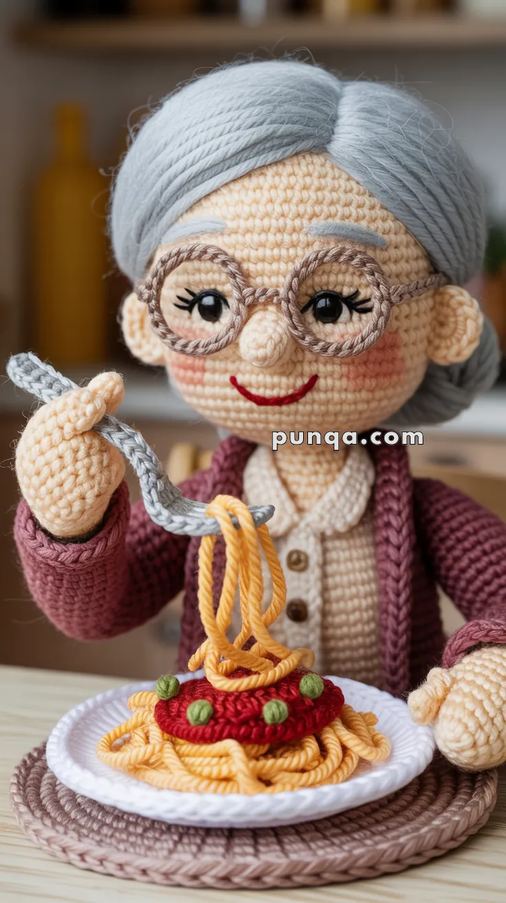 A crocheted figure of an elderly woman with glasses and gray hair, smiling while holding a fork with yarn spaghetti over a yarn plate of spaghetti with tomato sauce and peas.