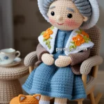 A crocheted doll of an elderly woman sitting in a rocking chair wearing a blue dress, brown shoes, and a white hat with orange flower accents. Beside her is a small crocheted orange cat and a book titled 'Knitting Patterns' on the floor. A teacup sits on a small crocheted table nearby.