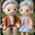 A crocheted elderly couple sits on a bench, holding hands. The man wears a brown jacket and beige pants, while the woman is dressed in a blue dress with a pink sweater. Both have gray hair and are smiling, with a background featuring a tree and flowers.