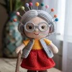 A crocheted doll resembling an elderly woman with gray hair, glasses, a yellow top, red skirt, and gray vest, holding a cane, and with colorful pins in her hair, placed on a wooden surface with a blurred floral background.