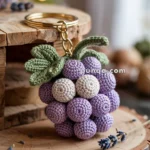 A crocheted keychain shaped like a bunch of grapes, with purple and cream-colored yarn, hanging on a wooden surface. The keychain includes green crocheted leaves and a gold metal ring.