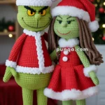 Two crocheted Grinch figures dressed in red Santa outfits with white trims and hats, standing together in a festive setting with blurred Christmas lights in the background.