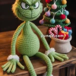 A crocheted green character with a mischievous grin sits on a wooden table next to a small crocheted Christmas tree and a basket of colorful yarn decorations.