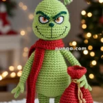 A crocheted green character with a mischievous smile, wearing a red scarf and holding a red sack, against a festive, blurred background with lights.