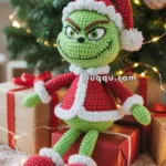 A crocheted Grinch doll dressed in a red Santa outfit sits on a wrapped gift in front of a decorated Christmas tree with lights and ornaments.