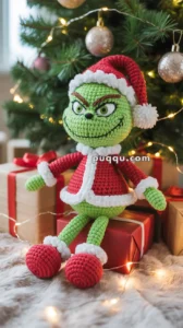 A crocheted Grinch doll dressed in a red Santa outfit sits on a wrapped gift in front of a decorated Christmas tree with lights and ornaments.