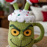 A knitted mug cozy resembling a green character with yellow eyes and a mischievous grin, featuring a fluffy white top with red decorations and two red and white striped socks sticking out.