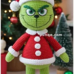 A crocheted green character dressed in a red Santa outfit, complete with a hat and boots, standing in front of a decorated Christmas tree with lights and ornaments.