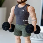 A crochet doll resembling a muscular man in a workout pose, wearing a dark tank top, green shorts, and holding black dumbbells. The doll features detailed stitching, brown hair, and a beard, with white and black sneakers.