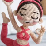 Crocheted doll in a yoga pose, wearing a pink outfit and headband, with a bun hairstyle, holding a pink ball and making an 'okay' gesture.