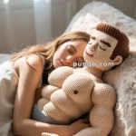 A woman sleeping in bed, cuddling a large, muscular crocheted doll. The scene is set in a softly lit room with a sheer curtain partially covering a window in the background.