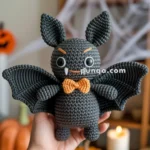 A handmade crocheted toy bat with large ears, a friendly expression, and an orange bow tie, held against a backdrop of a Halloween-themed room with pumpkins and candles.