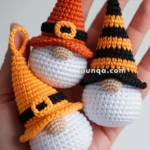Three crocheted gnome ornaments in Halloween colors, each with a white body and pointed hat, held in a person's hand. The hats are orange, yellow with a black belt, and orange with black stripes, all with embellishments resembling belts.